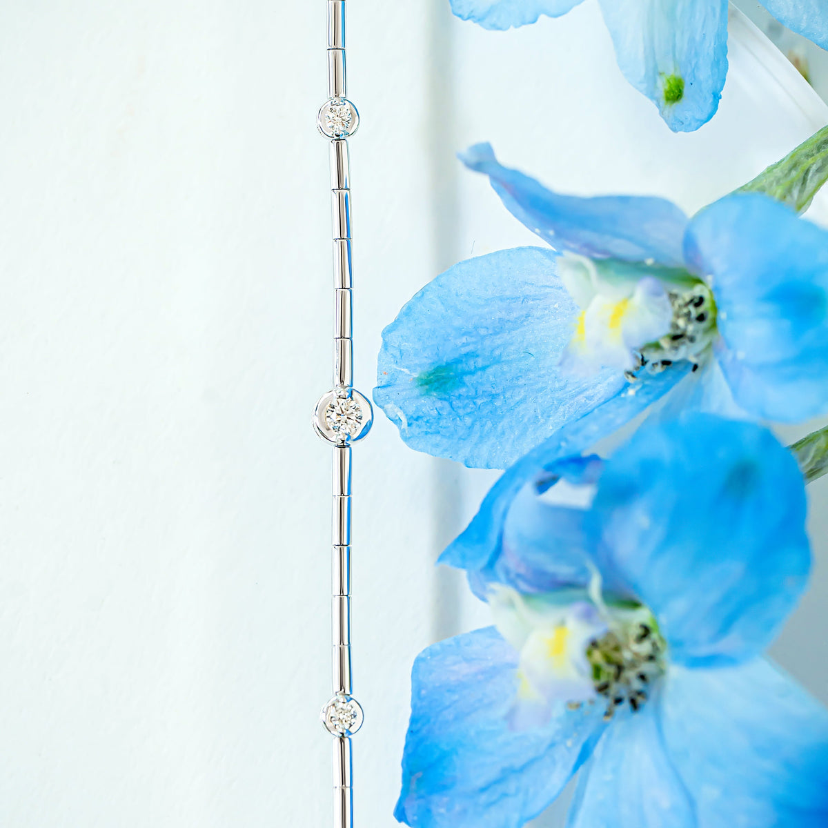 A close-up of a delicate white gold diamond station line bracelet, styled next to vibrant blue delphinium flowers against a soft white background.