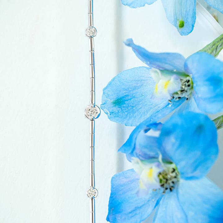 A close-up of a delicate white gold diamond station line bracelet, styled next to vibrant blue delphinium flowers against a soft white background.