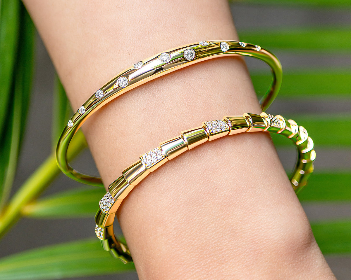 Two gold bangle bracelets. One with pavé-set diamonds on a serpent design, and another with embedded set diamonds, worn stacked on a person's wrist against a blurred green leaf background.