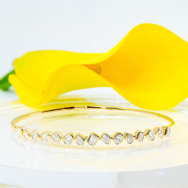 A gold bangle bracelet with a row of mixed-shape bezel-set diamonds, displayed on a clear acrylic stand with a bright yellow flower behind it against a white background.