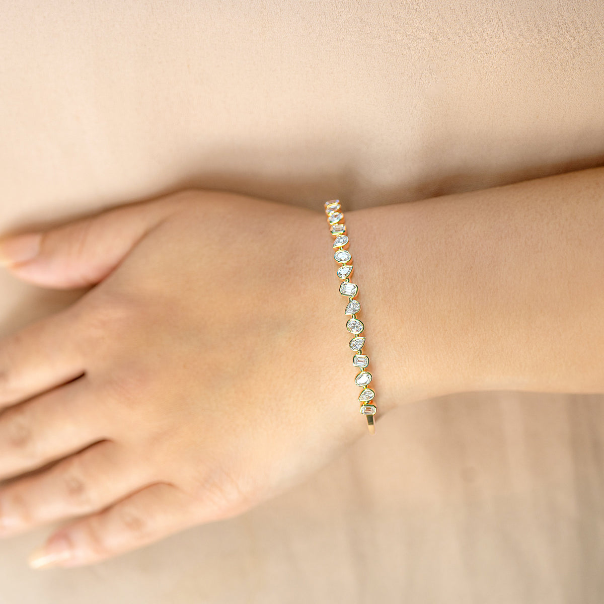 A close-up of a person's wrist wearing a delicate gold bracelet adorned with mixed fancy cut diamonds, set against a soft beige background.