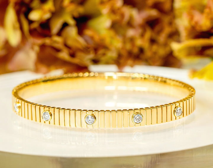 A gold fluted bangle bracelet with five round-cut diamonds, displayed on a white reflective surface with a blurred warm-toned background.