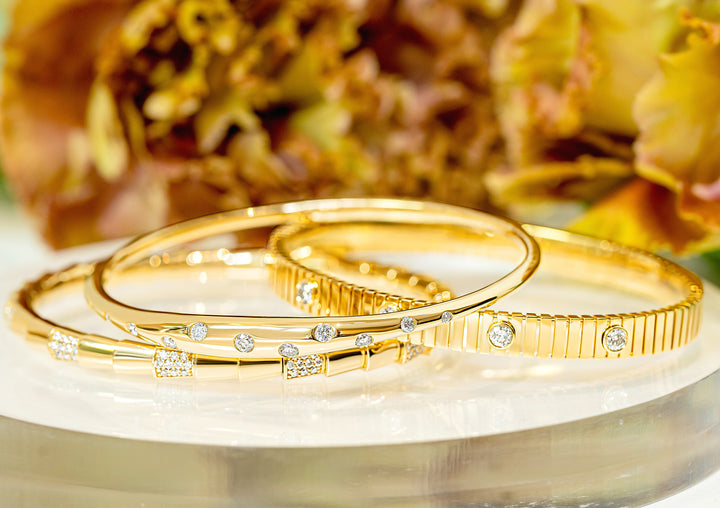 Three gold bangle bracelets with diamonds, displayed on a white surface with a blurred floral background.