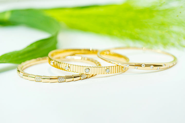 Three gold bangles with diamonds, arranged overlapping on a white surface with soft green blurred leaves in the background.