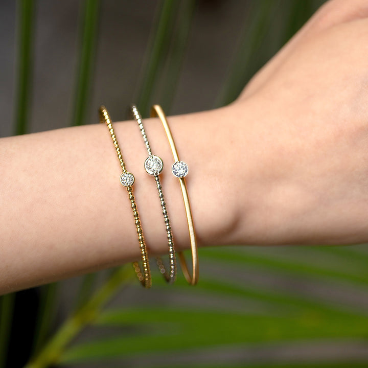 Three delicate flexible bangle bracelets worn on a person's wrist, featuring gold and silver metallic finishes with small round brilliant cut accent, set against a blurred green background.