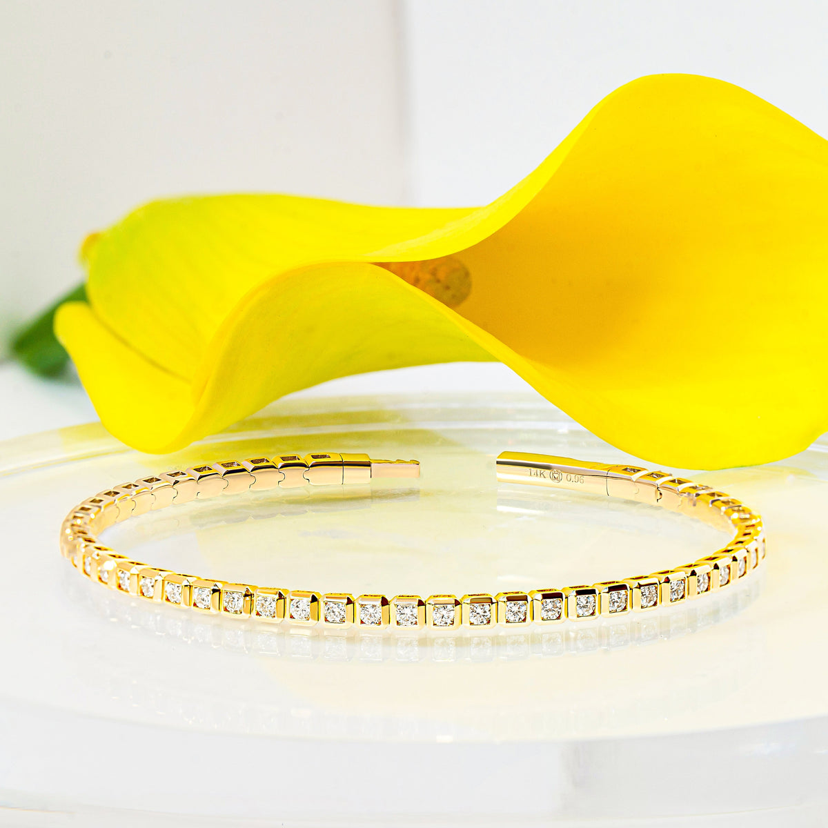 A gold bracelet with a row of diamonds, displayed on a clear acrylic stand with a bright yellow calla lily flower in the background.