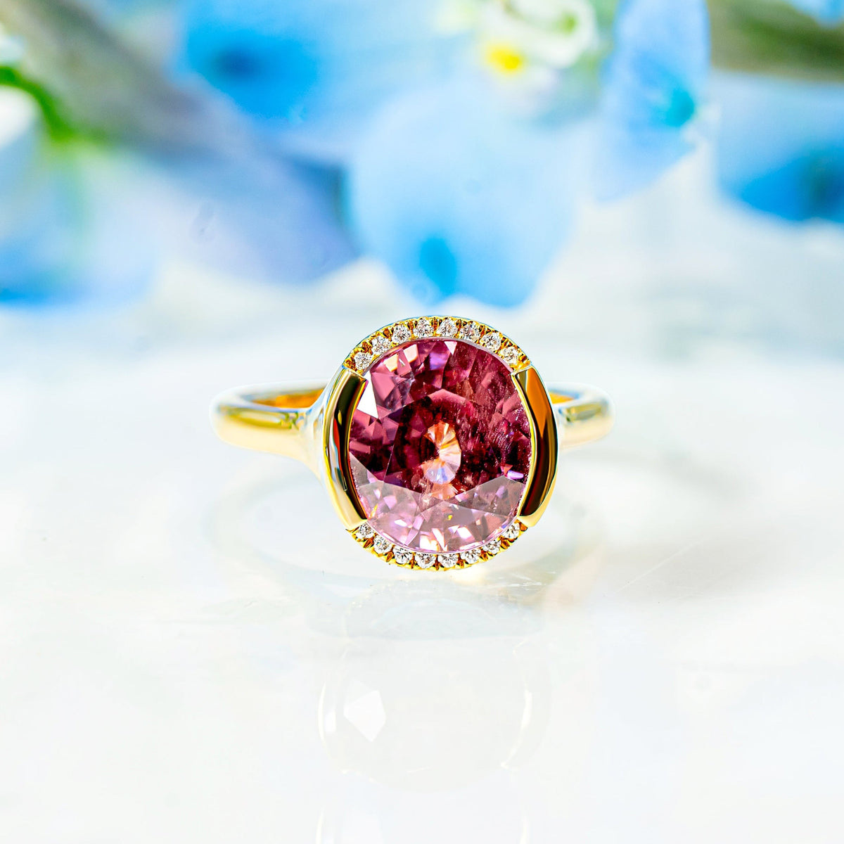A gold ring with a large oval pink zircon and a partial halo of small diamonds, resting on a white reflective surface with blurred blue flowers in the background.