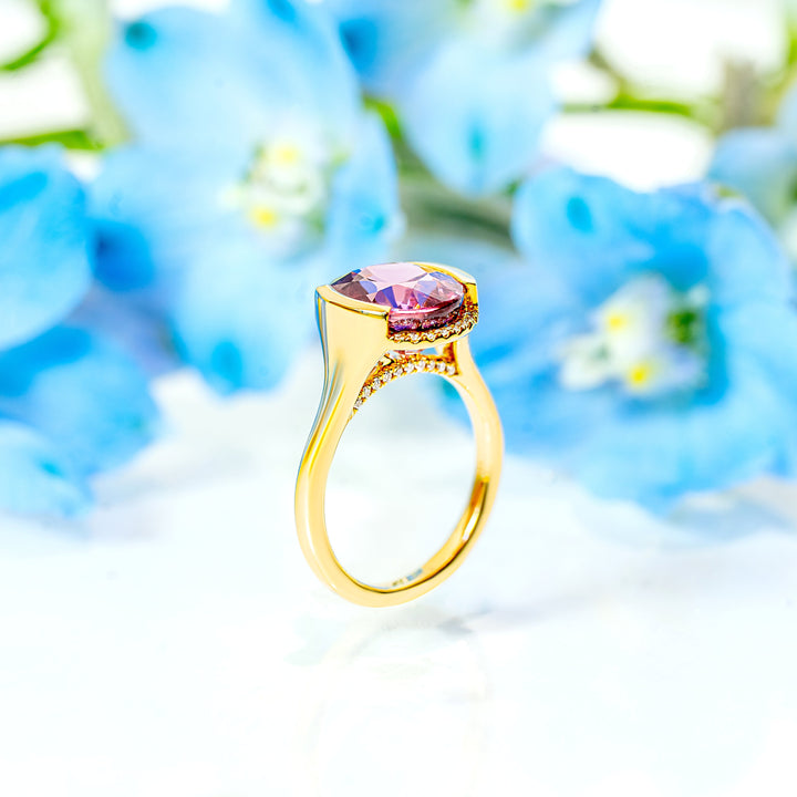A gold ring with an oval pink zircon and diamond accents on the bridge, styled on a white surface with blurred light blue flowers in the background.