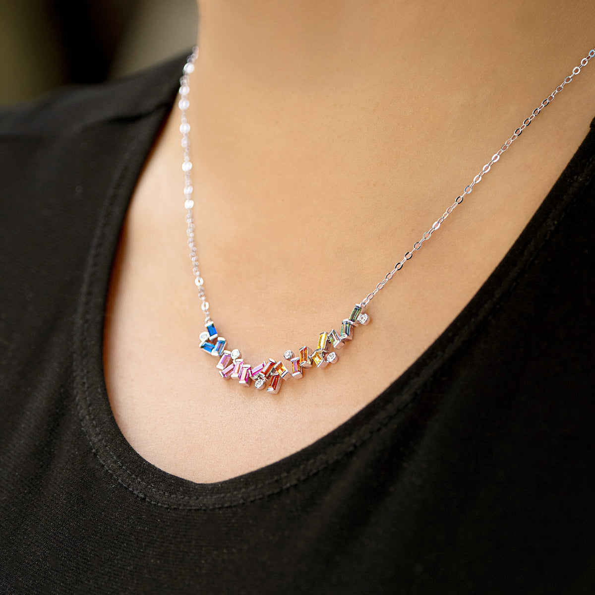 A close-up of a person wearing a delicate silver-tone chain necklace with a curved row of small, colorful gemstones in a rainbow hue, styled with a black top.