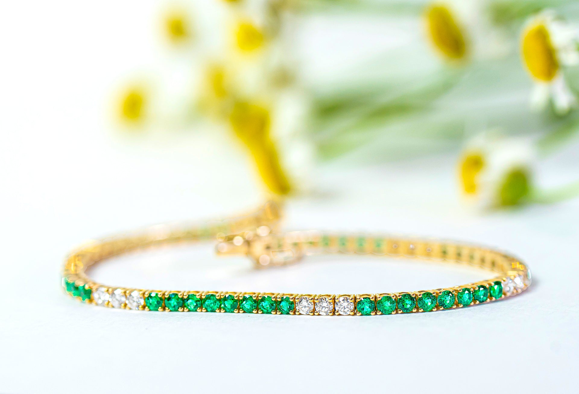 Gold tennis bracelet with green emeralds and diamonds on a white background with blurred flowers