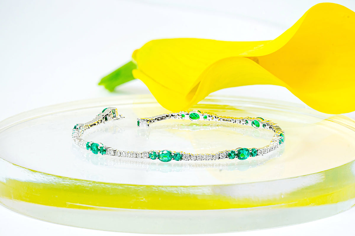 A white gold tennis bracelet with emeralds and diamonds, displayed on a clear acrylic riser with a yellow calla lily flower behind it against a white background.