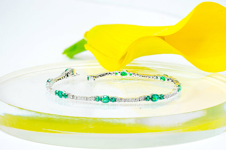 A white gold tennis bracelet with emeralds and diamonds, displayed on a clear acrylic riser with a yellow calla lily flower behind it against a white background.