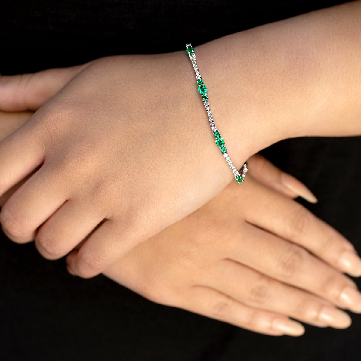 A close-up of a delicate bracelet worn on a person's wrist, featuring alternating emerald green gemstones and white diamonds set in a silver-toned white gold tennis style bracelet. The person's hands are crossed over, and they are wearing a black top.
