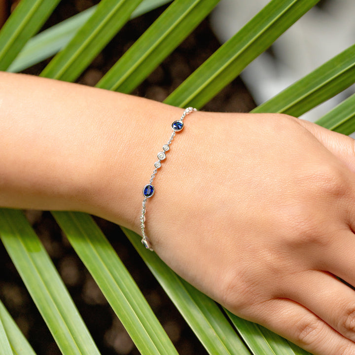 A delicate silver chain bracelet with small blue sapphires and white diamonds, worn on a person's wrist resting on green palm leaves.
