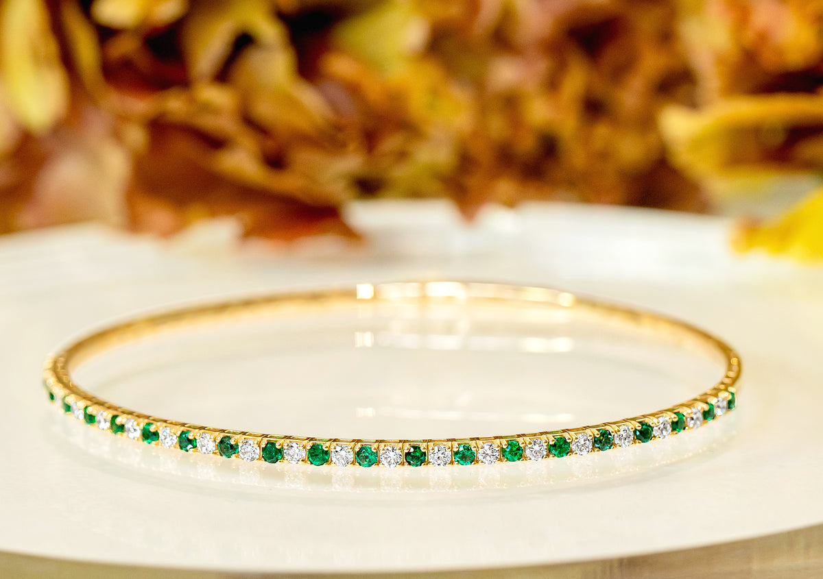 A gold bangle bracelet adorned with a row of round-cut green emeralds and small white diamonds, resting on a white surface with a soft-focus background of autumn leaves.
