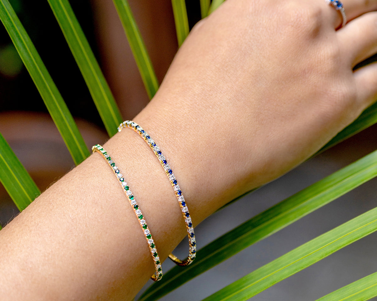 A close-up of a person's wrist wearing two delicate tennis-style bracelets, one with green stones and one with blue stones, both on a gold-tone band. A matching gold ring with a blue stone is also visible on the finger, set against a background of green palm leaves.
