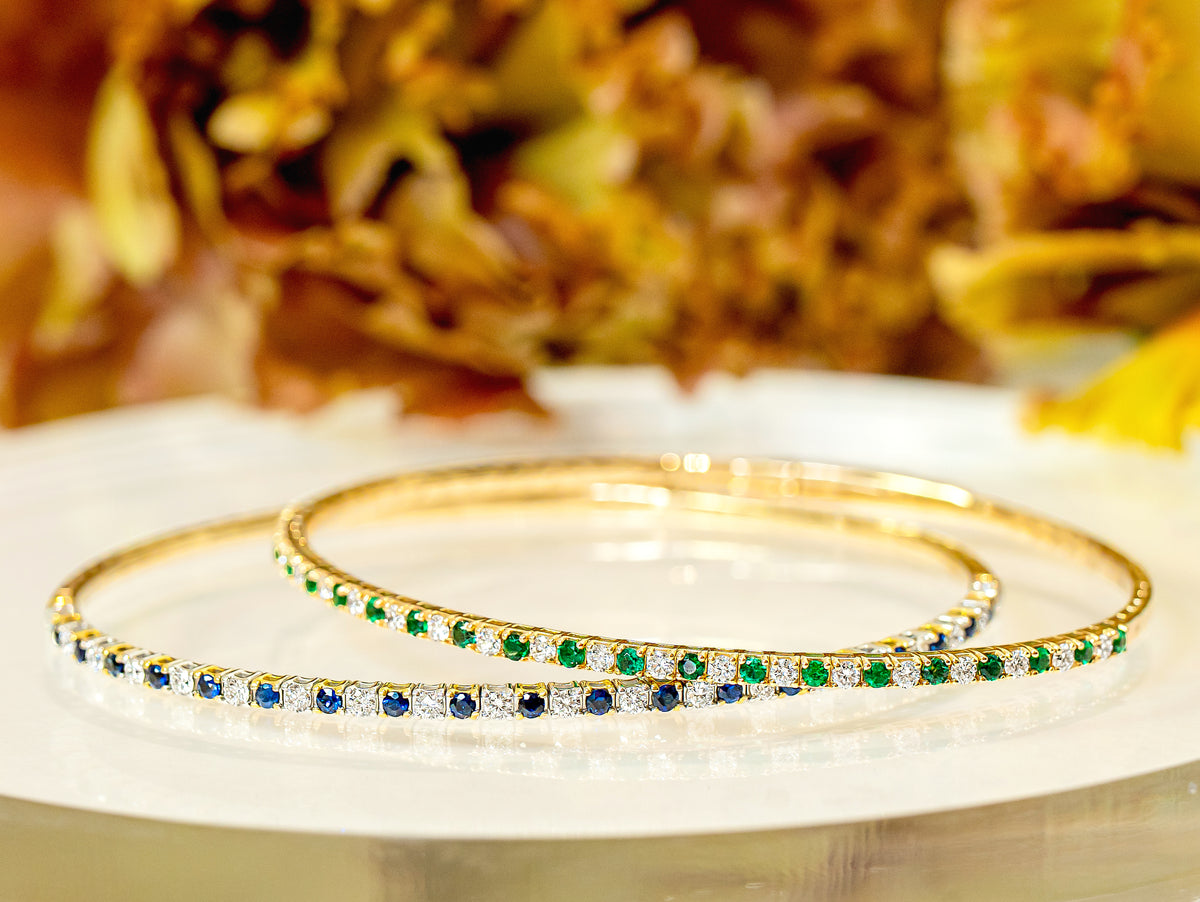 Two gold bangle bracelets with rows of green emeralds and blue sapphires, accented with small diamonds, displayed on a white surface with a warm, blurred background.
