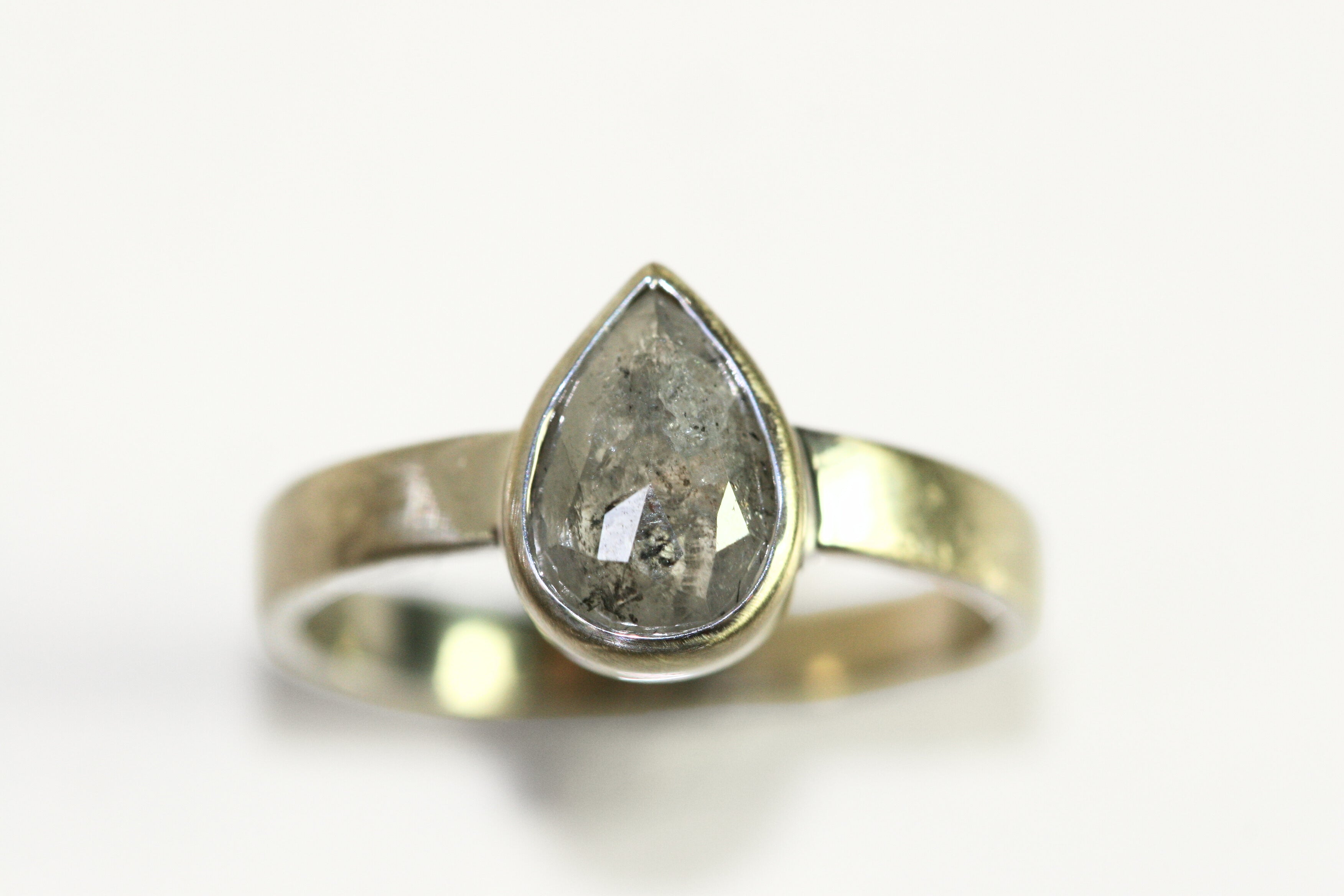 A pear shaped salt and pepper diamond is bezel set in white gold. The band is thick. The stone has interesting inclusions.