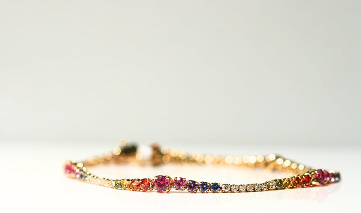 A bracelet made of yellow gold and rainbow of stones. The stones are white diamonds, and an array of green, yellow, orange, red, pink, purple, indigo, and blue sapphires. The stones are set in a rainbow and each is prong set.