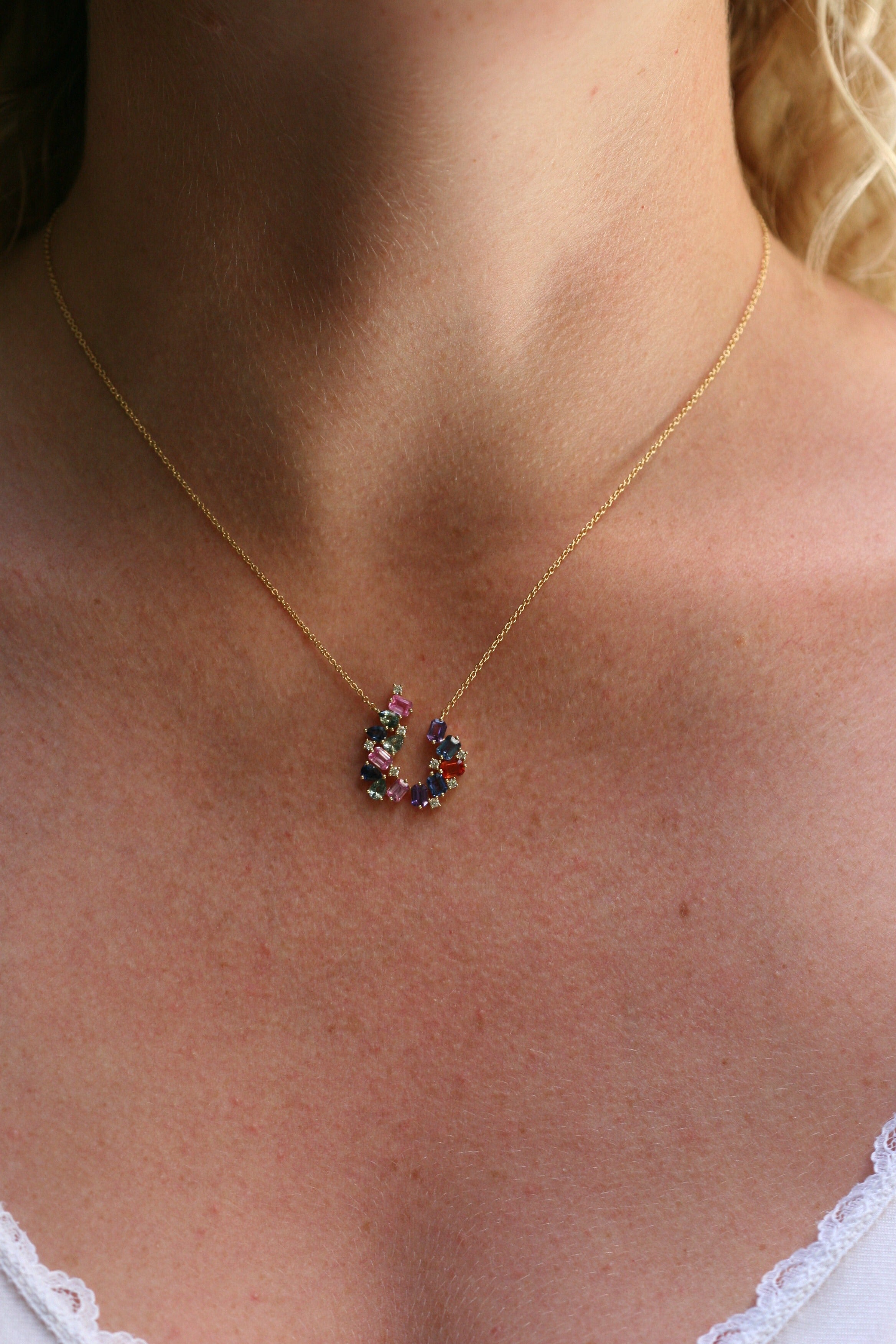 Pendant of multicolored gemstones hangs off a yellow gold chain. The stones are different shapes - emerald cut, pear cut, and round diamonds. The stones are blue, green, orange, pink, and purple. The stones are arranged in an open circle. This photo shows the necklace on a model. The necklace ends an inch below her collarbones. The chain is dainty.