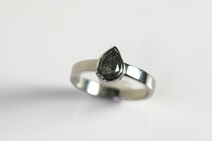 A pear shaped salt and pepper diamond is bezel set in white gold. The band is thick. The stone has interesting inclusions.