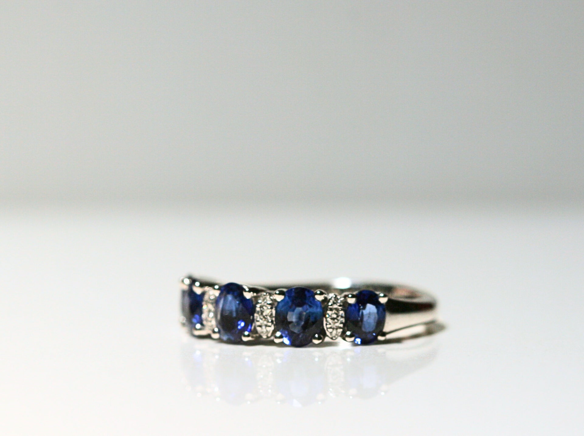 A 3/4 view of a white gold ring that features sapphires and diamonds. There are 5 deep blue, oval cut sapphires. Between each are two white diamonds, that are set in white gold and look like marquise shapes. The band of the ring is solid white gold.