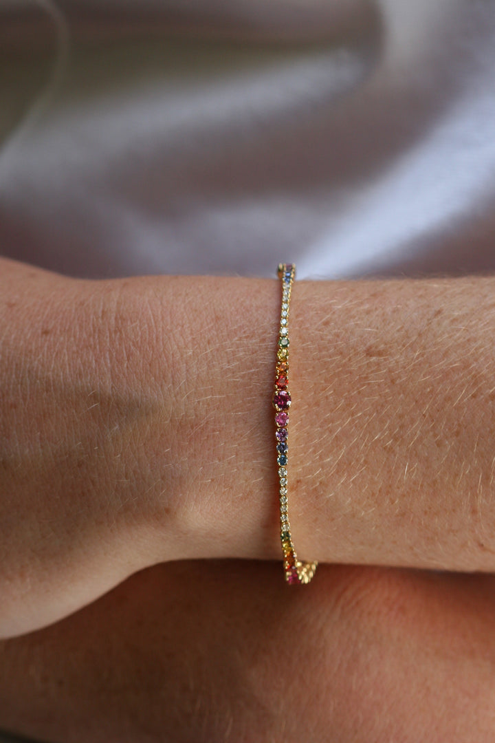 This shows the bracelet on a lady's wrist. A bracelet made of yellow gold and rainbow of stones. The stones are white diamonds, and an array of green, yellow, orange, red, pink, purple, indigo, and blue sapphires. The stones are set in a rainbow.