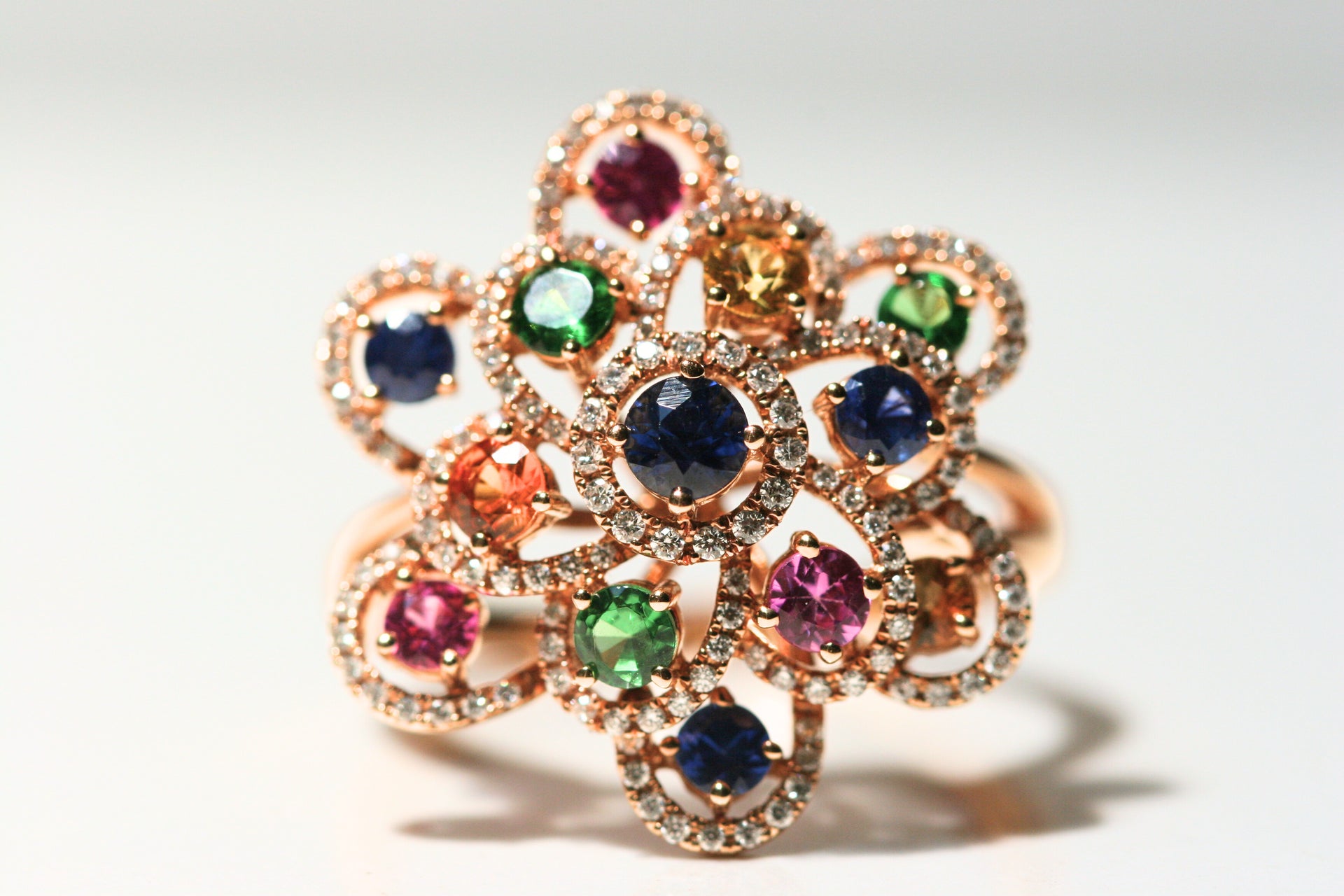 Cocktail flower ring. Open petals have a single stone floating in the middle. Stones are blue, green, yellow, orange, pink, and all are set in rose gold. The outline of each petal is set with white round diamonds. The design is whimsical and unique.