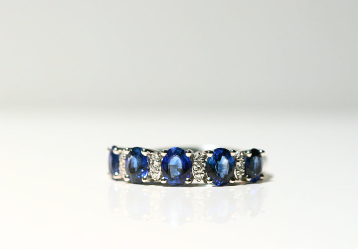 A white gold ring that features sapphires and diamonds. There are 5 deep blue, oval cut sapphires. Between each are two white diamonds, that are set in white gold and look like marquise shapes. The band of the ring is solid white gold.