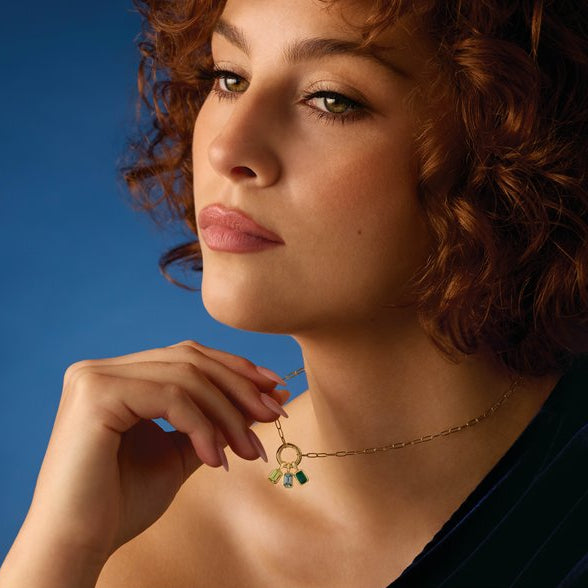 Woman wearing a gold necklace with emerald, blue zircon, and peridot charms against a blue background