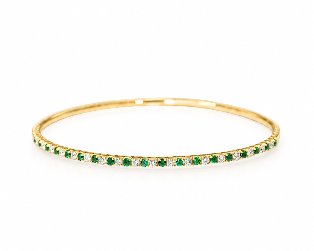 A thin yellow gold bangle bracelet featuring a single row of round-cut green emeralds and diamonds, shown on a plain white background.