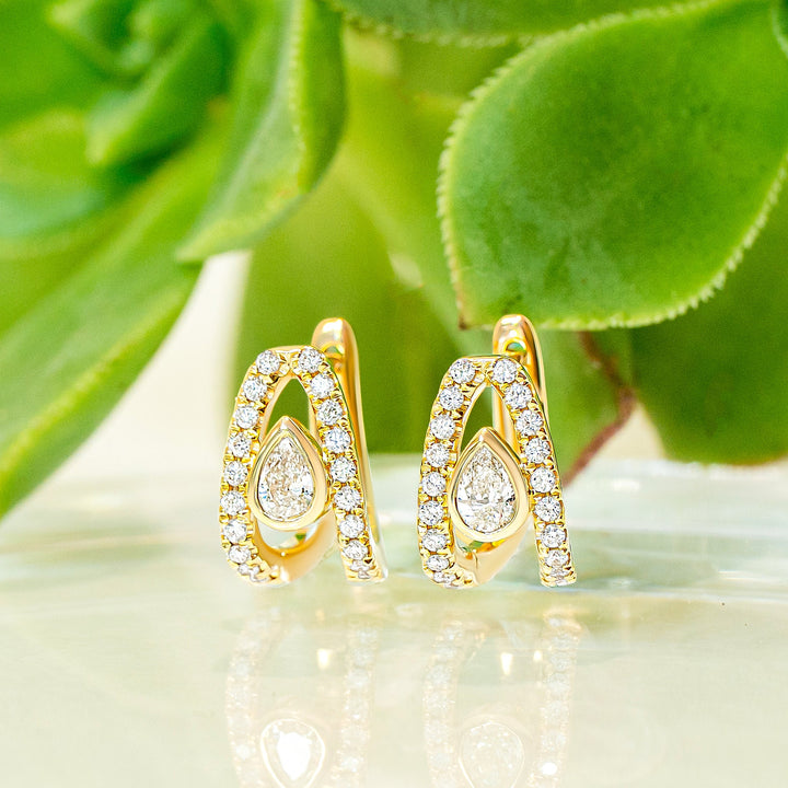 A pair of gold huggie hoop earrings with teardrop-shaped diamond, displayed on a reflective white surface with green succulent leaves in the background.